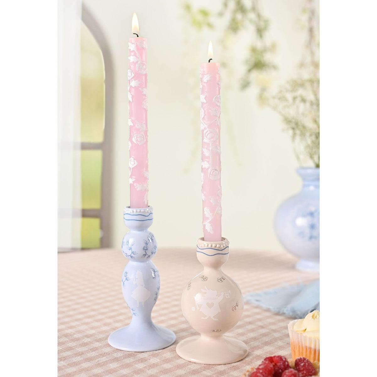 Set of two ceramic taper candle holders, Alice in Wonderland design, with pink floral candles lit, on a dining table setting.