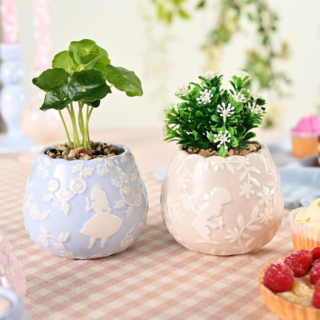Set of 2 mini round ceramic planters with scalloped rims, featuring Alice in Wonderland relief designs, one blue holding a leafy plant, one beige holding white flowers.