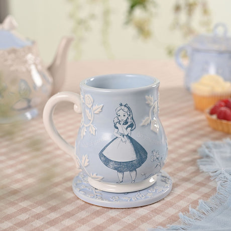 Blue embossed earthenware mug with Alice in Wonderland sketched in blue-grey lines, raised white roses and script detailing, shown on a matching coaster with teapots in the background.