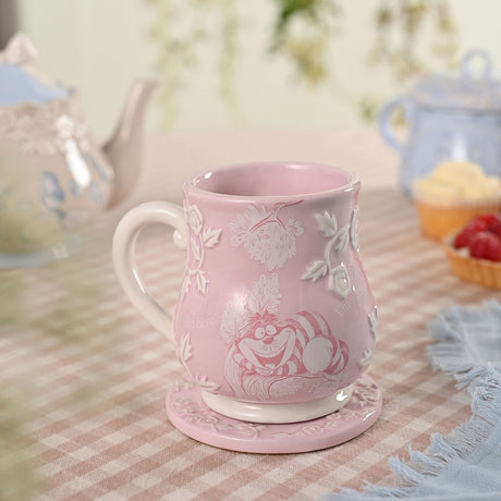 Pink earthenware mug with embossed white roses and leaves, featuring a rose-line Cheshire Cat design and script text, shown with a matching coaster on a tea table.