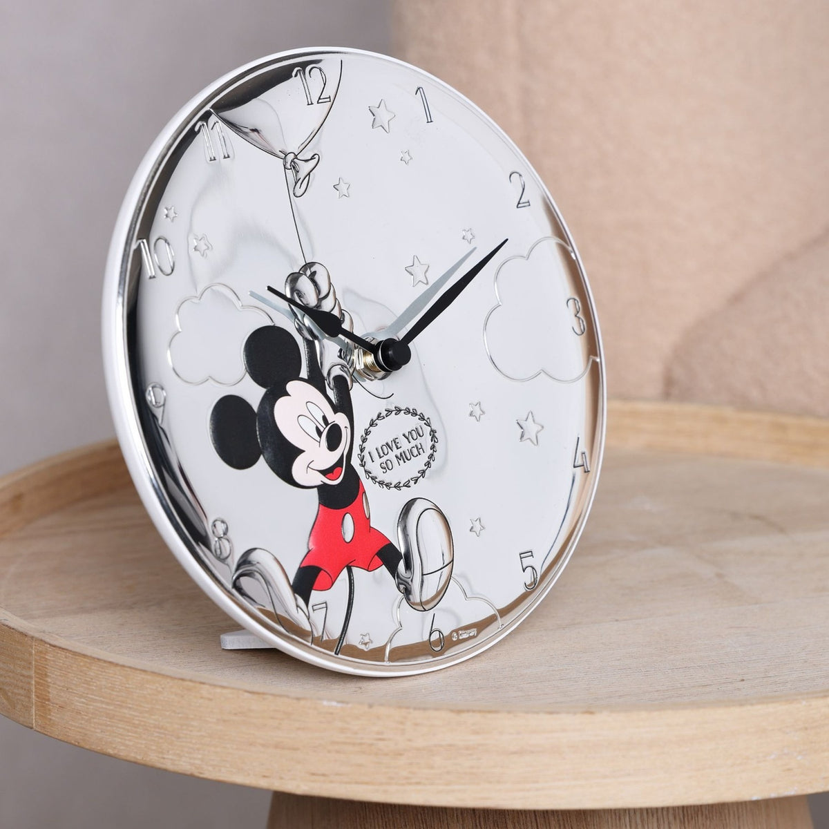 Round silver-plated wall clock with Mickey Mouse in red shorts holding a balloon, embossed clouds and stars, displayed on a wooden table.
