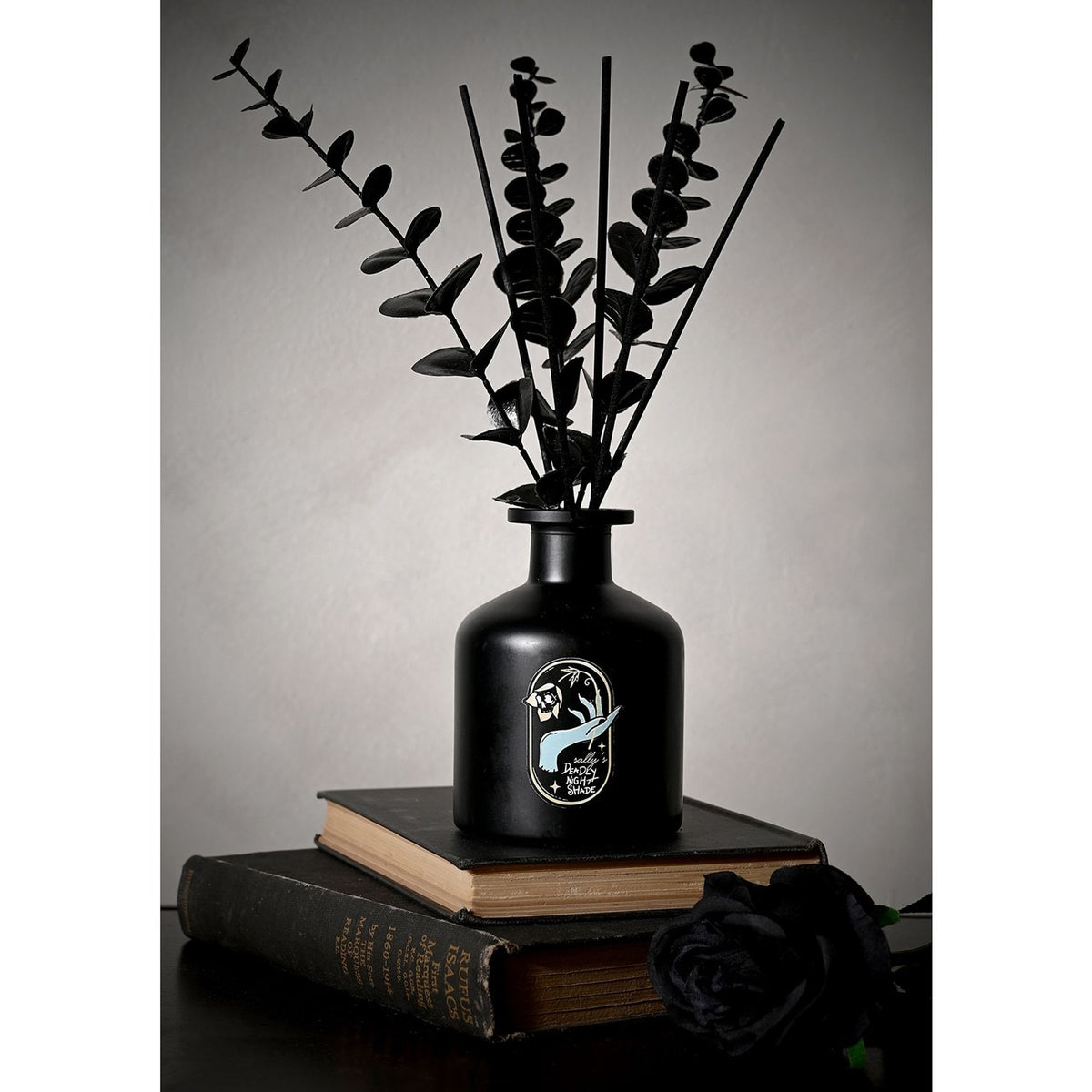 Black reed diffuser bottle with black leafy stems, featuring a gold-edged label of a pale blue hand holding a bloom, set on stacked books against a grey background.