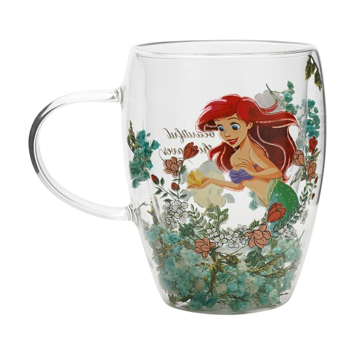 Clear glass mug with a printed design of Ariel the mermaid holding a shell amid pink and white flowers, dried green florals layered at the base, on a white background.