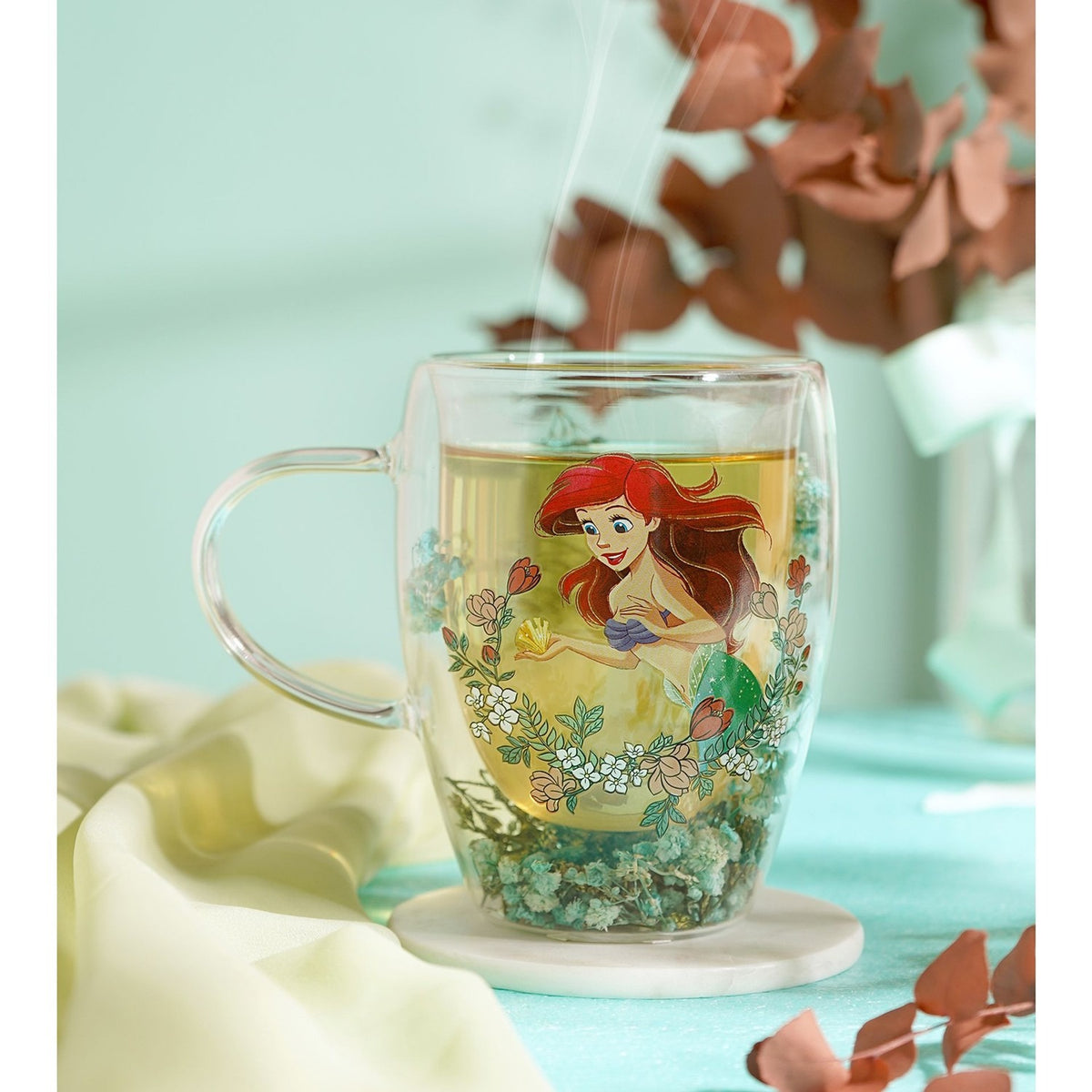 Double-walled glass mug with clear handle, featuring Ariel the mermaid and floral wreath design, filled with tea and dried green botanicals at the base, shown steaming on a marble coaster.