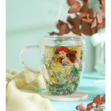 Double-walled glass mug with clear handle, featuring Ariel the mermaid and floral wreath design, filled with tea and dried green botanicals at the base, shown steaming on a marble coaster.
