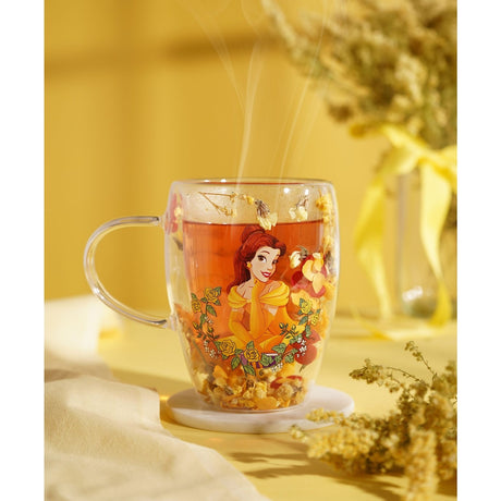 Glass mug with an image of Belle in a yellow gown encircled by yellow roses, filled with hot tea and dried yellow and orange flowers, set on a marble coaster.