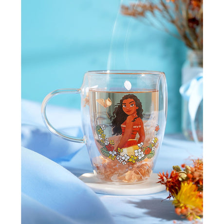 Clear glass mug with a Moana illustration encircled by red, yellow and white flowers, filled with hot tea and dried petals, set on a light surface.