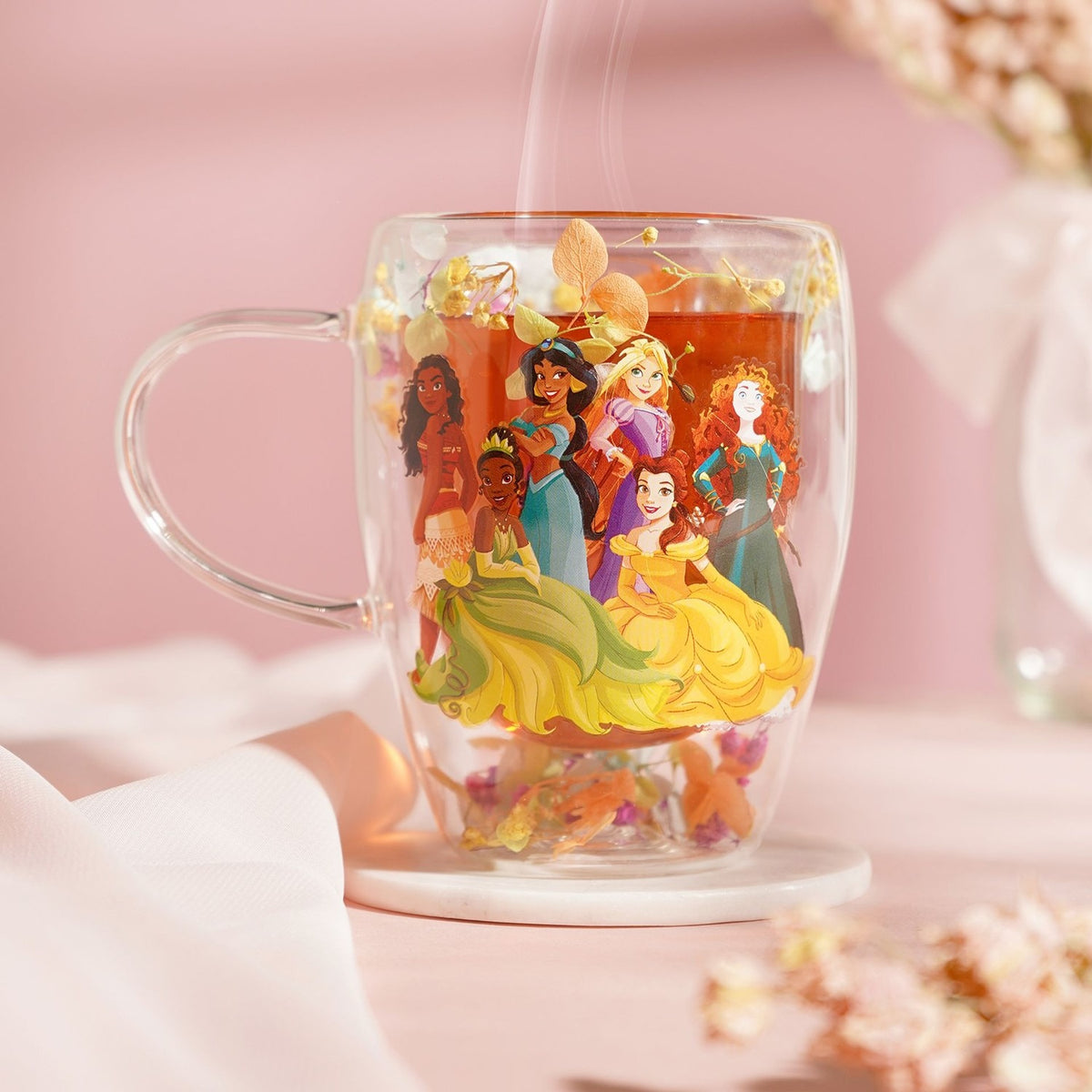 Double-walled glass mug, 320ml, with a multicoloured Disney princess decal, filled with tea and set on a marble coaster amid dried flowers on a pink background.