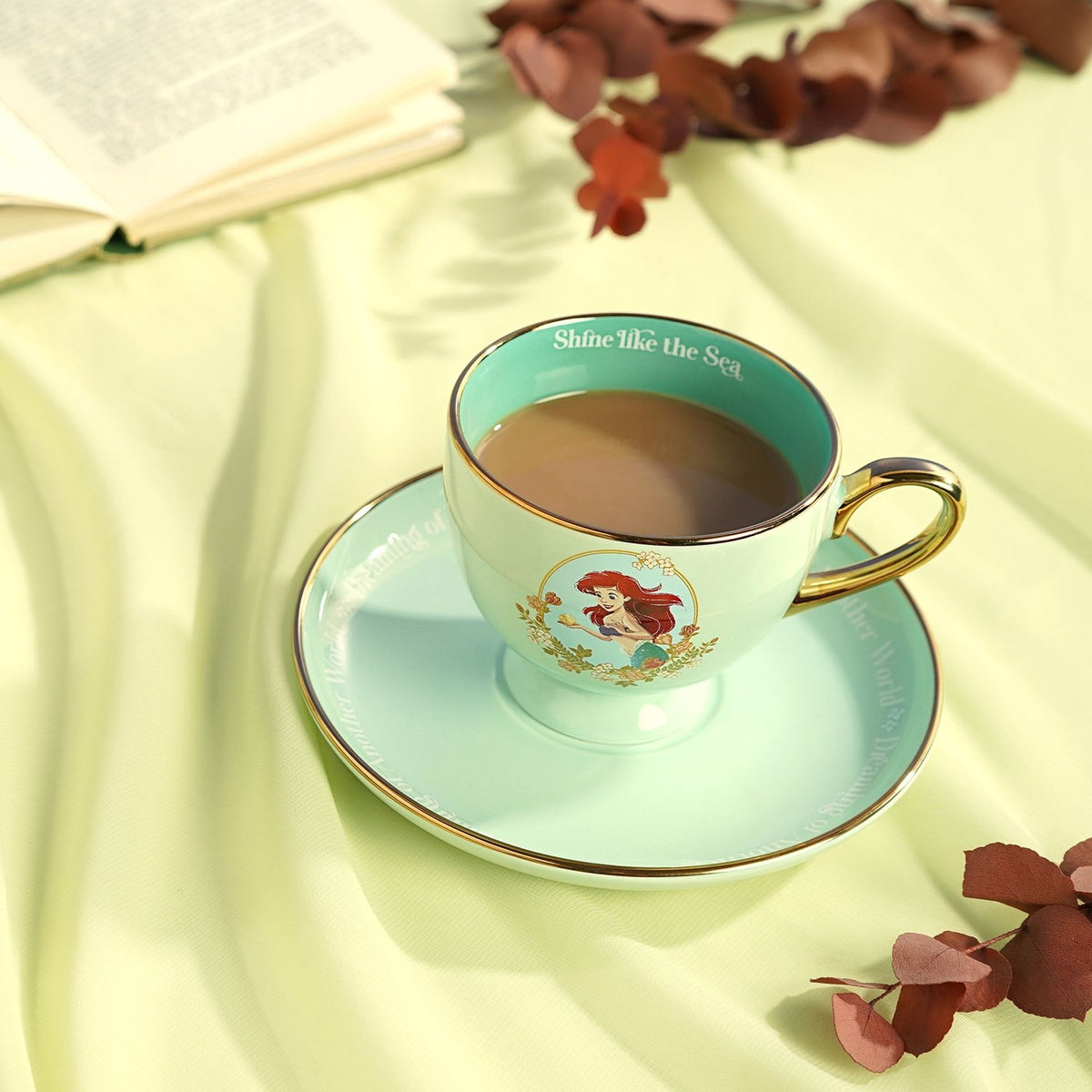 Pastel green porcelain tea cup and saucer with gold handle and rim, printed Ariel decal, "Shine like the Sea" text inside, filled with tea on a fabric surface.