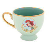 Pastel green porcelain tea cup with gold handle and rim, footed base, and a decal of Ariel holding a shell framed by floral garland, deeper green glaze inside, on a white background.