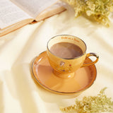 Yellow porcelain footed tea cup and matching saucer with a Belle illustration and "Bold as a Rose" script inside the rim, shown filled with tea on a cream fabric surface.