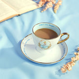 White porcelain tea cup and saucer with gold trim, filled with tea, featuring a Cinderella medallion and "Follow your dreams" text, styled with an open book and pink flowers on blue cloth.