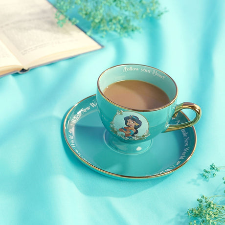 Green porcelain tea cup and saucer with gold rim, featuring a Princess Jasmine portrait, filled with tea on a teal fabric surface beside an open book.