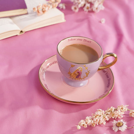 Rapunzel Pastel Princess Porcelain Tea Cup & Saucer in lilac and pink with gold rim, handle and footed base, filled with tea, styled on a pink fabric surface with a book and dried flowers.