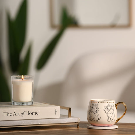 White porcelain Sketched Marie Kitten Mug with grey pencil-style cat sketches, gold rim, handle and lettering, pink base, shown beside a candle and book on a table.