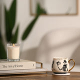 Jack & Sally Porcelain Mug 400ml in cream with black sketched artwork, gold lettering and glossy gold handle, shown on a coaster beside a candle and book on a wooden table.