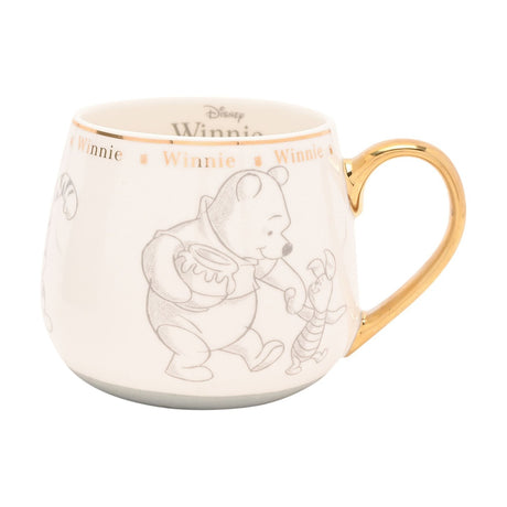 White porcelain Winnie the Pooh mug with gold handle and rim, featuring grey sketched illustrations of Pooh and Piglet, gold "Winnie" lettering band, on a white background.