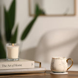 White porcelain Tinker Bell mug with grey pencil-style sketch and gold handle, shown on a coaster beside a lit candle and book on a wooden table.