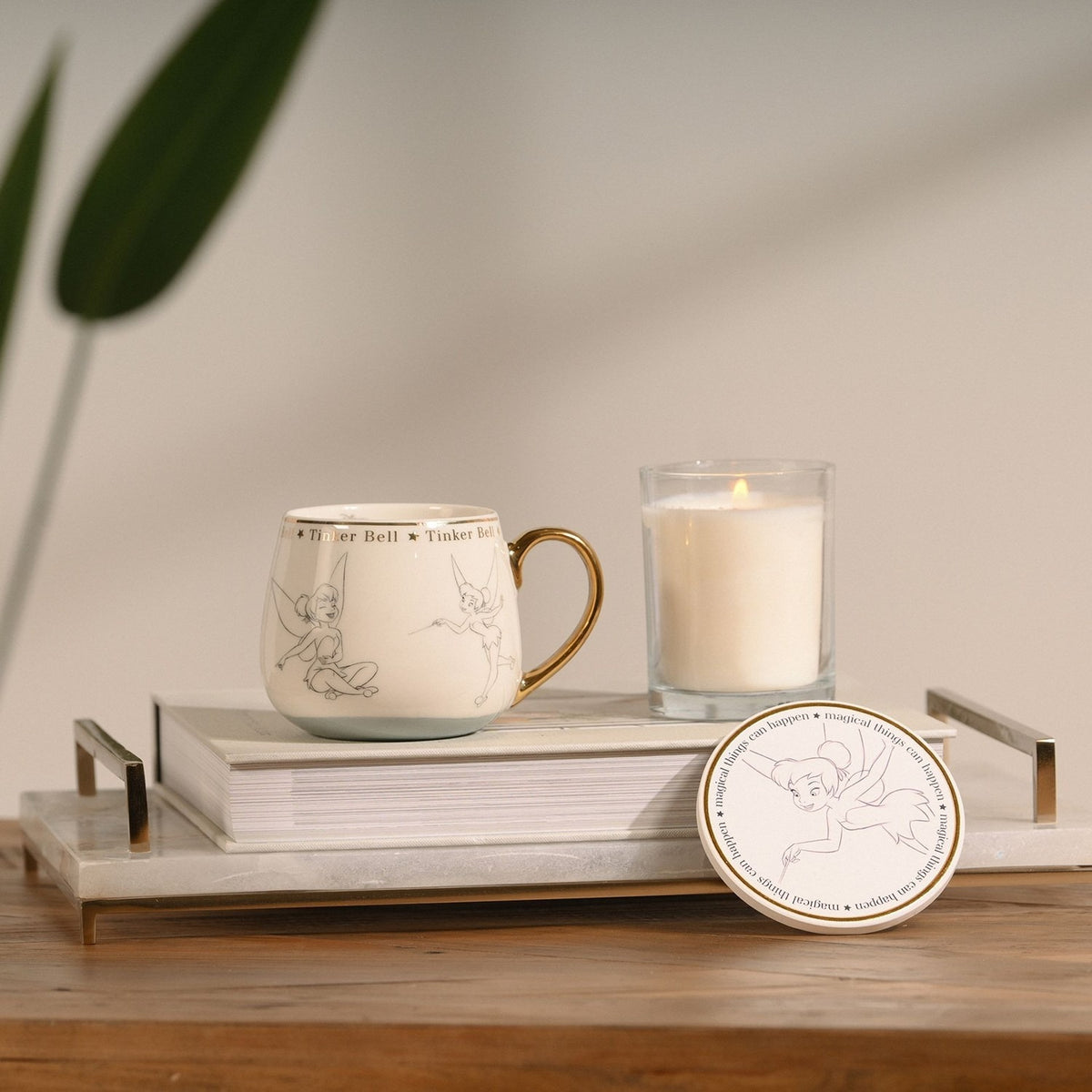 Tinker Bell Classic Collectable Porcelain Mug in white with gold handle and rim band, shown beside a matching coaster and lit candle on a marble tray in a home setting.