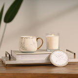 Tinker Bell Classic Collectable Porcelain Mug in white with gold handle and rim band, shown beside a matching coaster and lit candle on a marble tray in a home setting.