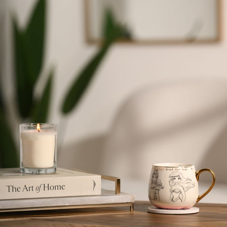 White porcelain Princess Classic Collectable mug with sketched grey princess portraits, gold rim and handle, pale pink base, displayed on a coaster beside a candle and book.