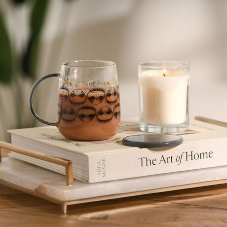 Clear glass Nightmare Before Christmas mug with grey handle, printed with repeating Jack Skellington faces, filled with hot chocolate beside a lit candle and books on a wooden table.