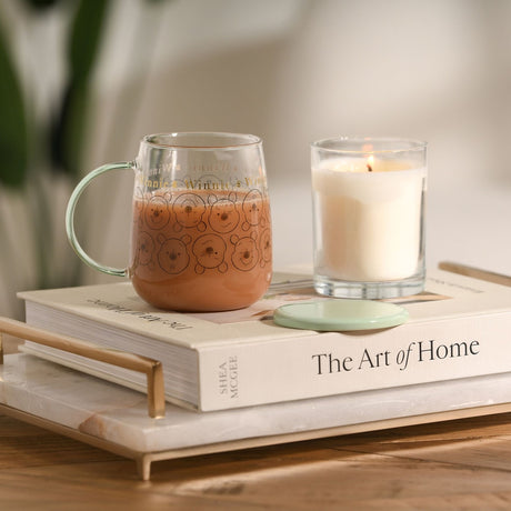 Winnie the Pooh Face Print Glass Mug with Lid 570ml in green, filled with a drink, shown on a book with a lit candle beside it on a tray.