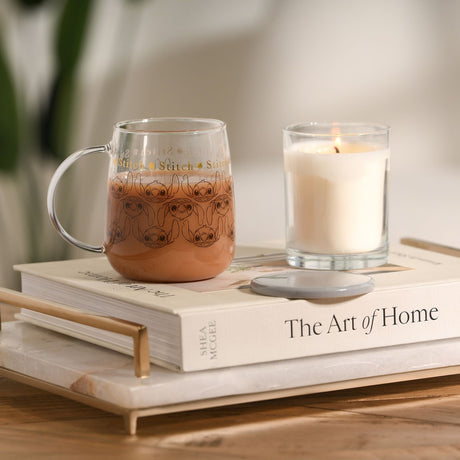 Glass mug with grey lid, filled with hot chocolate, showing Stitch's face repeated in grey line art under gold lettering, resting on a book beside a lit candle.
