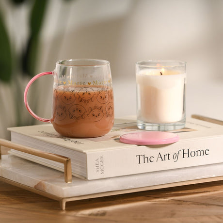 Marie Repeat Cat Face Print Glass Mug with Lid 570ml in pink, filled with a hot drink, pink handle and lid resting beside it, shown with a candle on a book in a home setting.