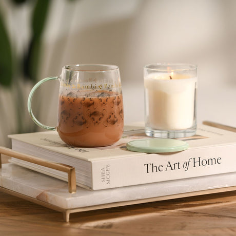 Bambi Sketch and Gold Lettering Glass Mug with Lid in green, filled with a drink, shown beside a lit candle on books in a home setting.