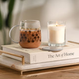 Mickey Mouse Classic Collectable Glass Mug with Lid in Grey, clear glass body printed with sketch-style Mickey faces and gold lettering, shown filled with a drink beside a lit candle on a book.