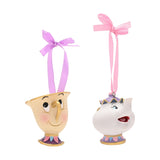 Chip and Mrs Potts resin hanging Christmas tree decorations, cream teacup and white teapot with painted faces, gold rims, purple and pink ribbons, on a plain white background.