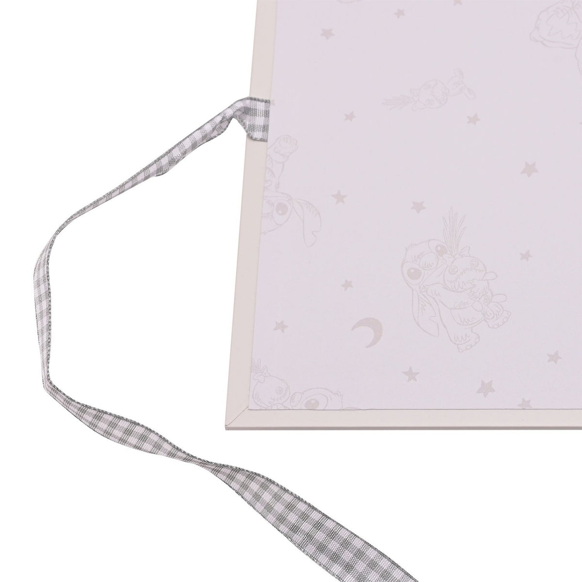 Close-up of the Ohana Stitch Baby Photo Album's corner, showing a cream cover with faint pastel character and star motifs, tied with a grey gingham ribbon.