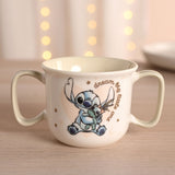 Cream porcelain baby mug with two sage-green handles, featuring Stitch hugging a green rag doll and "dream big little one" script, set against a softly lit background.