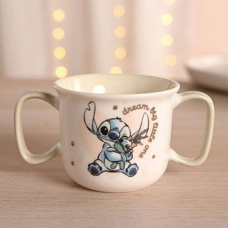 Cream porcelain baby mug with two sage-green handles, featuring Stitch hugging a green rag doll and "dream big little one" script, set against a softly lit background.