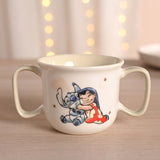 Cream porcelain mug with two sage-green handles, featuring Lilo hugging Stitch among stars and moons, 150ml capacity, shown on wood with bokeh lights behind.