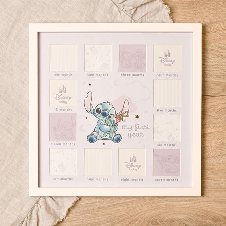 White wooden multi-aperture photo frame with twelve square month-labelled openings around a central Stitch design, pale mount, clouds and stars, on a neutral fabric and wood surface.