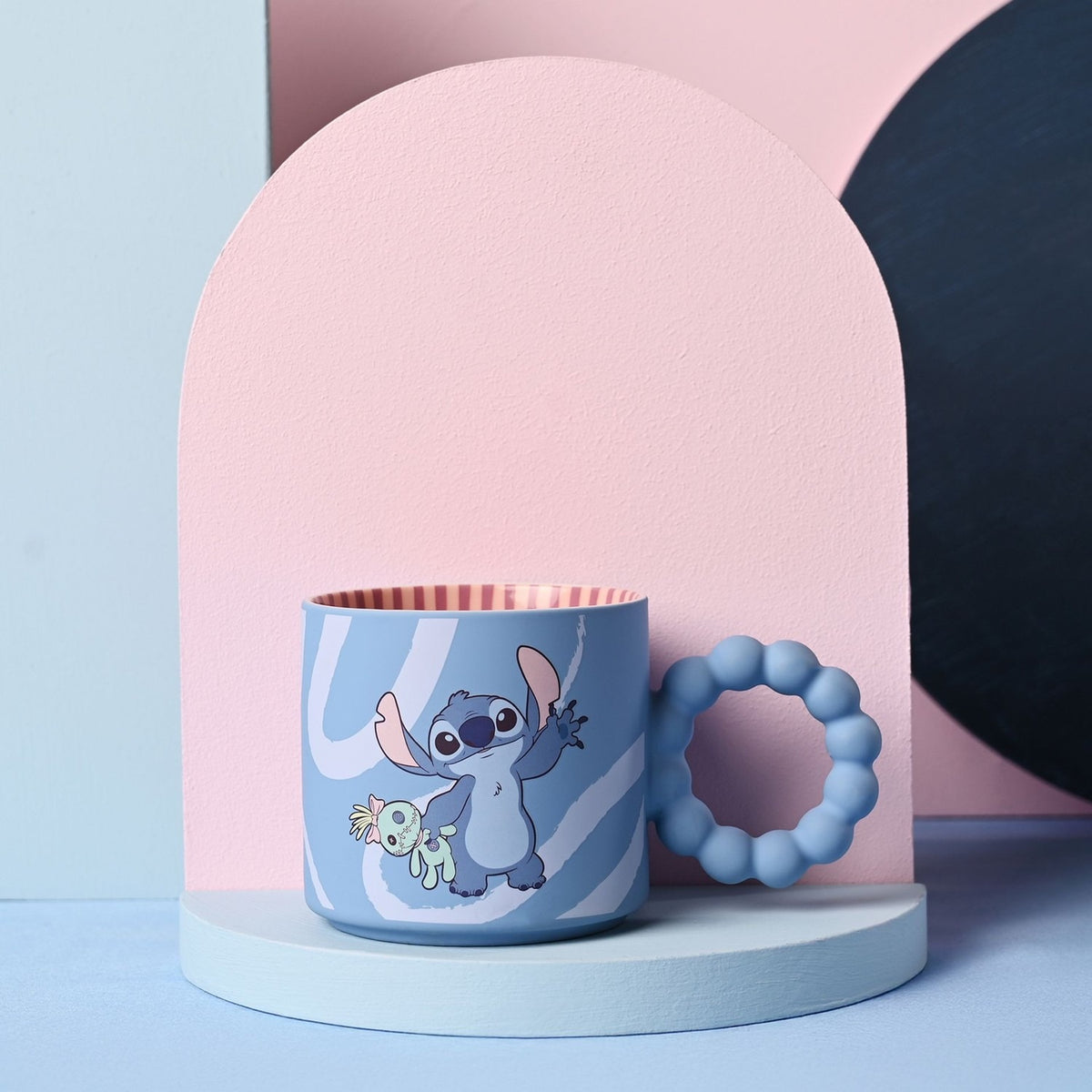 Blue ceramic Stitch Swirl mug with chunky bobble handle, white brushstroke swirls, Stitch waving with a green rag doll, red-striped interior, shown on a stand against a pink arch backdrop.