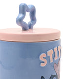 Close-up of the Stitch Stoneware Storage Jar, blue ceramic round body with raised pale pink "STITCH" lettering, pale pink lid and wavy star-shaped knob, on a white background.