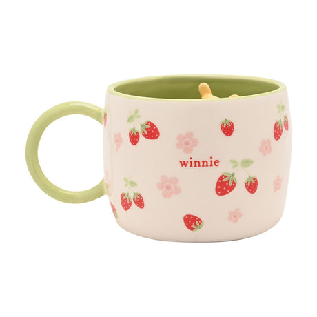 Cream stoneware mug with green handle and rim, printed with red strawberries, pink blossoms, and the word "winnie", featuring a 3D Winnie the Pooh figure inside, on a white background.