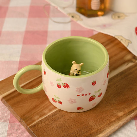 Winnie the Pooh Strawberry 3D Stoneware Mug in cream with green interior, featuring a 3D Pooh figure inside and red strawberries with pink blossoms outside, displayed on a wooden board.