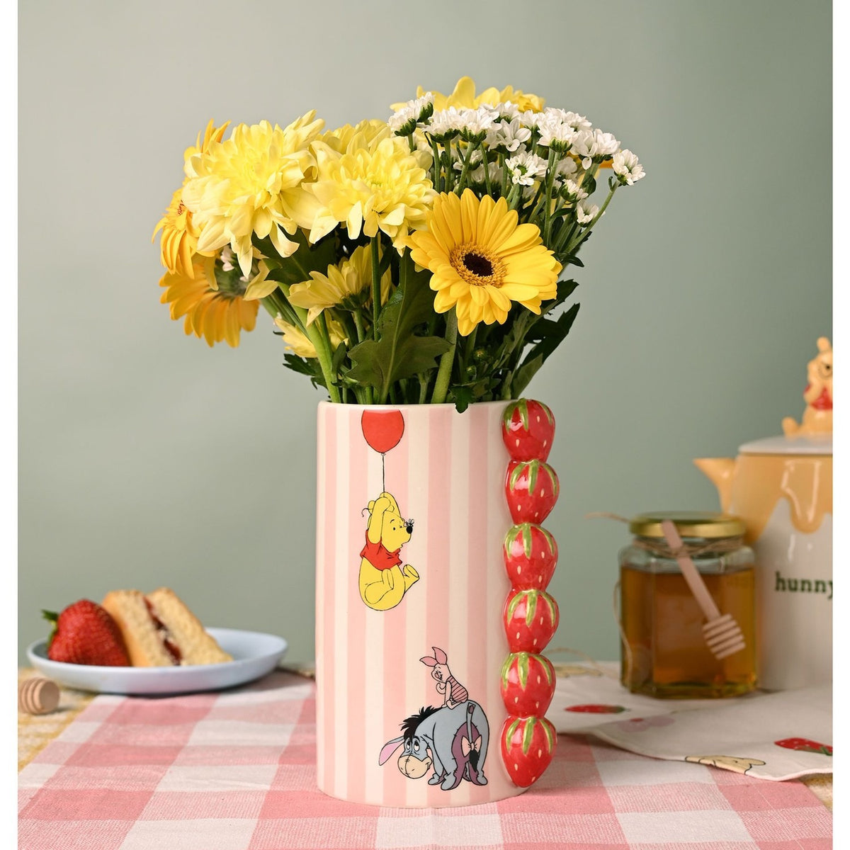 Winnie the Pooh Strawberry Stoneware Vase in pink and cream stripes with raised red strawberry column, Pooh and balloon design, holding yellow and white flowers on a picnic table setting.