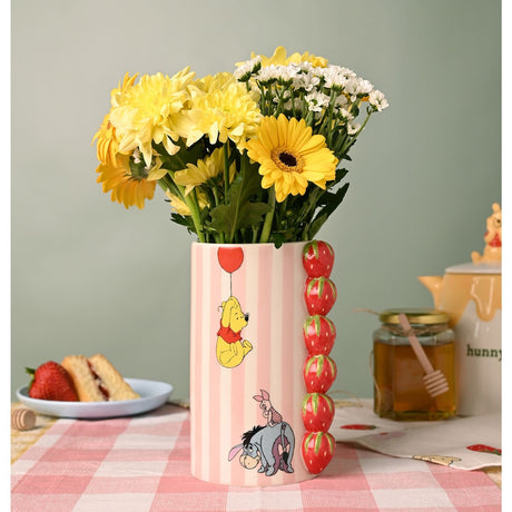 Winnie the Pooh Strawberry Stoneware Vase in pink and cream stripes with raised red strawberry column, Pooh and balloon design, holding yellow and white flowers on a picnic table setting.