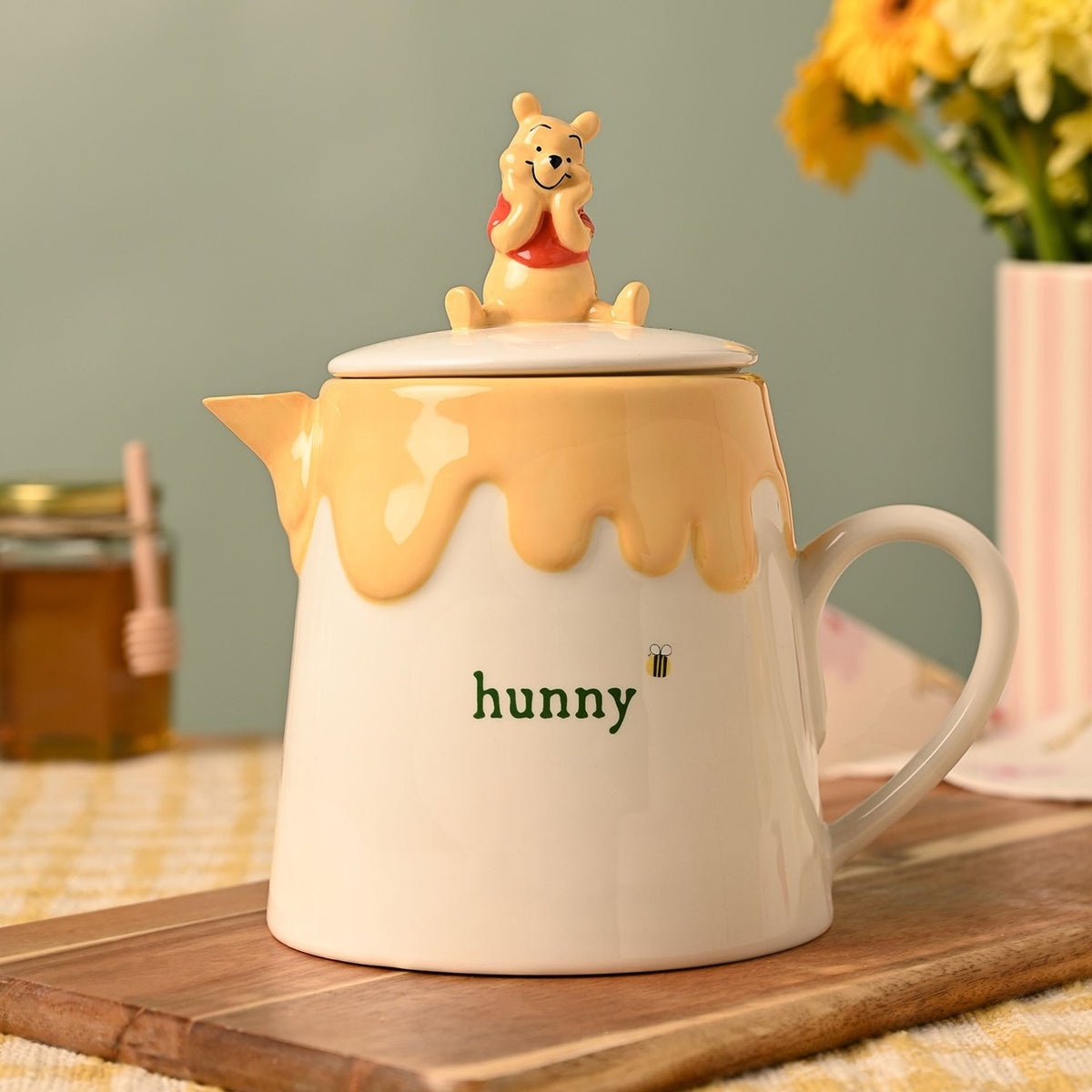 Winnie the Pooh Honey Pot stoneware teapot in cream and yellow, with dripping glaze, "hunny" text and bee design, topped with a Pooh figure lid, shown with flowers in the background.