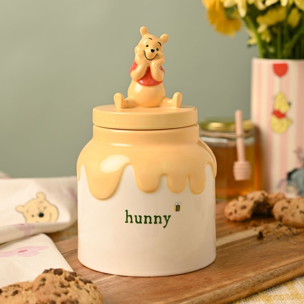 Ceramic Winnie the Pooh honey pot storage jar with yellow drip design and "hunny" lettering, shown on a kitchen table with cookies and flowers.
