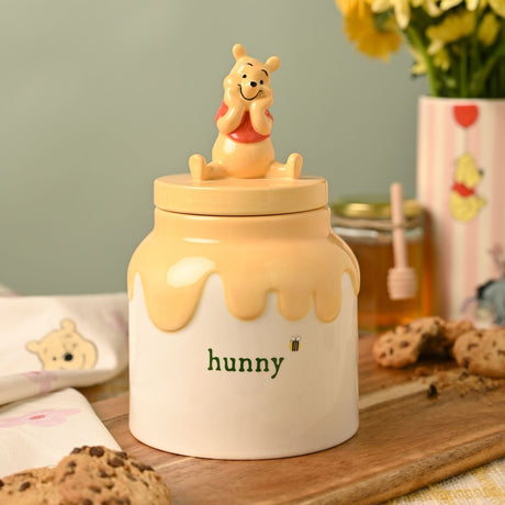 Ceramic Winnie the Pooh honey pot storage jar with yellow drip design and "hunny" lettering, shown on a kitchen table with cookies and flowers.