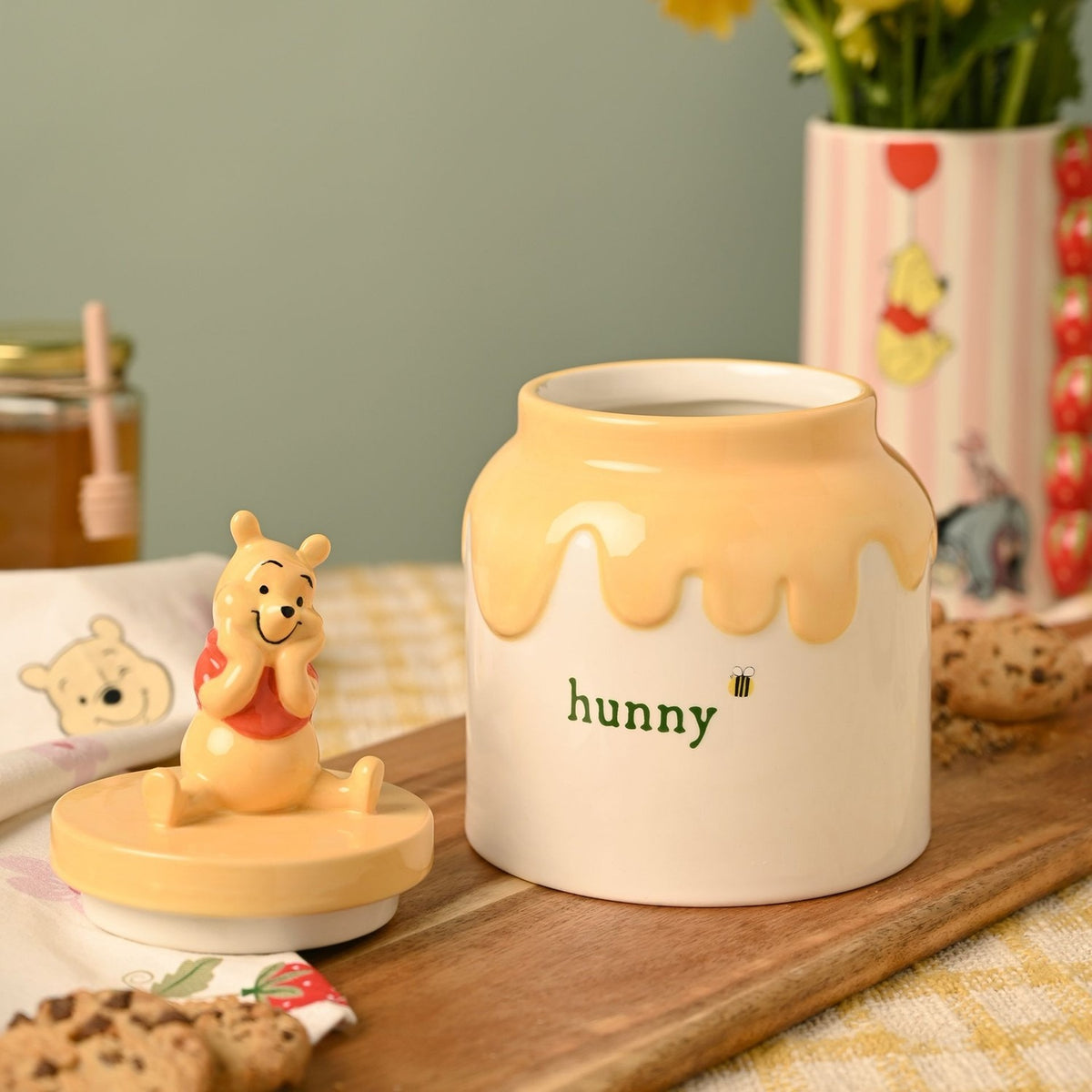 Cream stoneware honey pot with yellow drip finish, "hunny" lettering, bee design, and a seated Pooh figure on the lid, shown on a wooden board with cookies and honey.