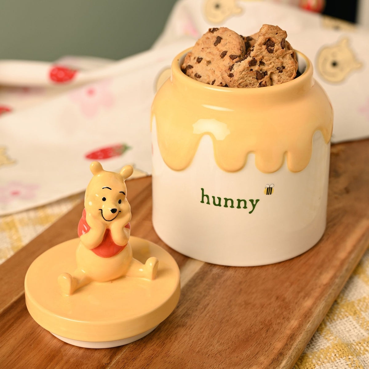 Winnie the Pooh Honey Pot stoneware storage jar in cream and yellow with a drip-effect rim and "hunny" lettering, filled with biscuits, lid with seated Pooh figure beside it on a wooden board.