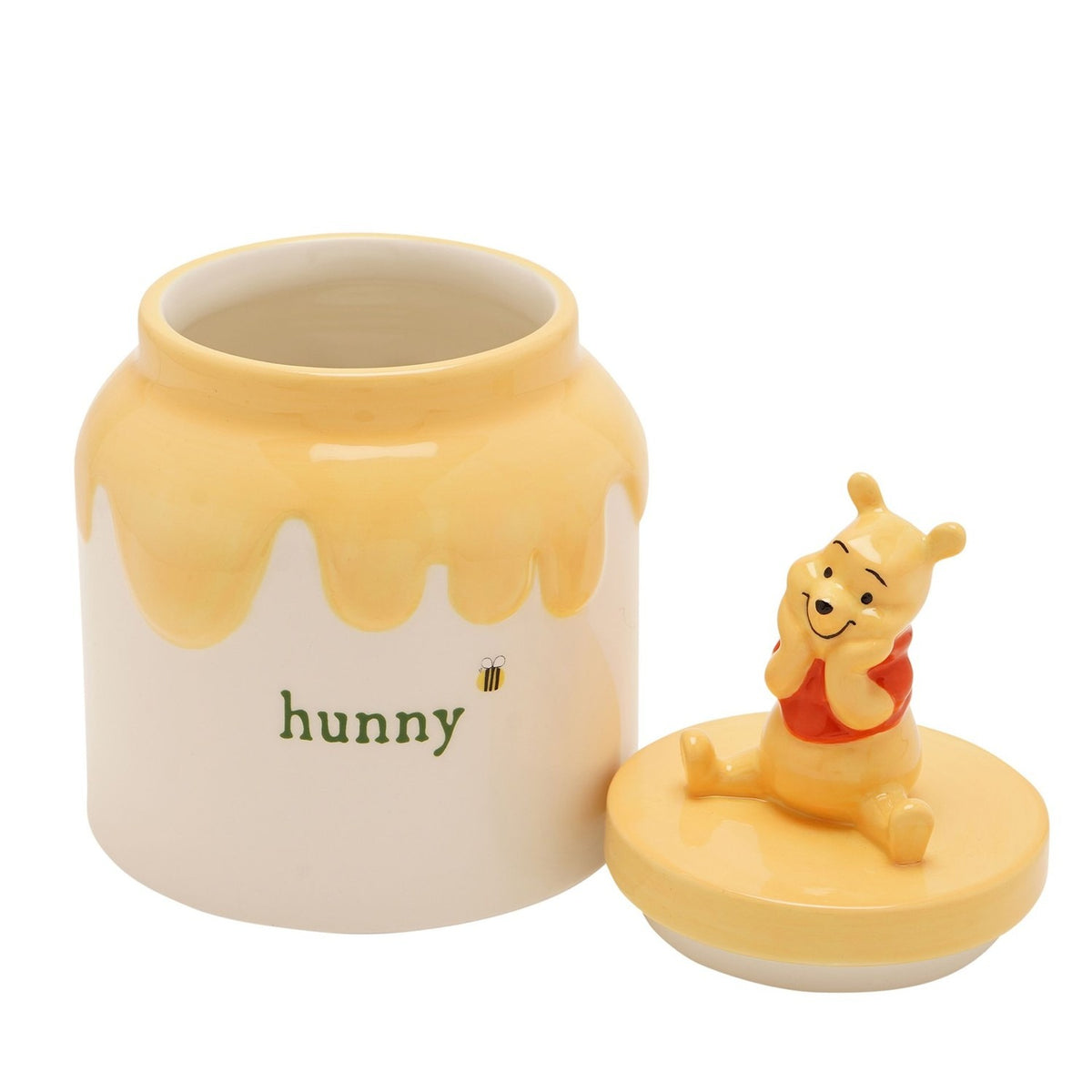 Ceramic honey pot storage jar in cream with a yellow drip effect, "hunny" text and bee detail, lid featuring a seated Winnie the Pooh figure, on a white background.