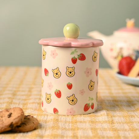 Winnie the Pooh Flower Stoneware Storage Jar in cream and pink, with a scalloped flower-shaped lid and green ball knob, decorated with Pooh faces, strawberries and blossoms, shown on a kitchen table.